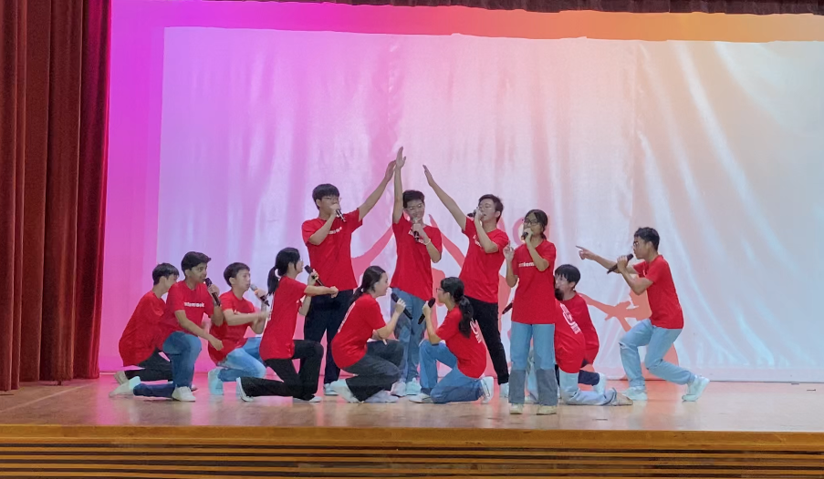 A group of teens in red shirts perform on stage, some kneeling and others standing with microphones and raised hands.